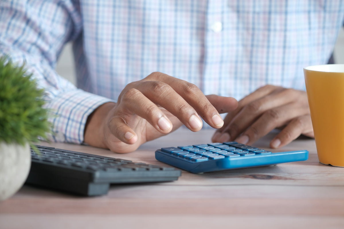 Un hombre con camisa a cuadros utiliza una calculadora azul sobre un escritorio de madera, al lado de un teclado negro y una taza amarilla.